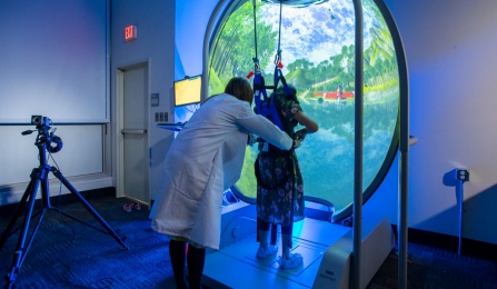 Faculty researcher positions a participant in a harness and platform to measure balance, in front of a 5-foot convex virtual reality screen. 