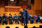 A PhD student wearing blue graduation regalia receives his hood from his faculty advisor on stage. 
