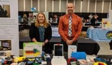 Image of two people standing in front of a table with a variety of assistive technology devices on display. 
