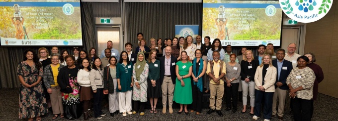 Group photo of approximately 30 attendees at the FoodWISE Asia Pacific Regional Meeting in September 2025. The participants are standing in rows inside a modern conference room with neutral beige walls, carpeted flooring, and bright overhead lighting. Two large screens in the background display event branding with an image of a person carrying water and the FoodWISE Asia Pacific logo. Attendees are dressed in business casual attire, including dresses, blazers, button-down shirts, and slacks. The overall color palette features earthy tones with pops of green and blue from clothing and the event logo, creating a professional yet welcoming atmosphere. At the bottom of the image, logos for UNSW Sydney, University of Technology Sydney, Northwestern University, and UNSW Global Water Institute are visible. 