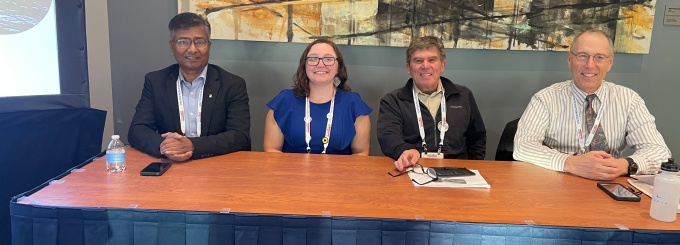 Four individuals seated behind a long rectangular table with a wooden top and dark blue table skirt at a conference setting. Items on the table include a water bottle, smartphone, notebook, and a large white cup. All participants wear lanyards, suggesting a formal event or panel discussion. Behind them is a large painting featuring abstract barn-like structures in earthy tones of orange, brown, and black. A projection screen is partially visible on the left side of the image. 