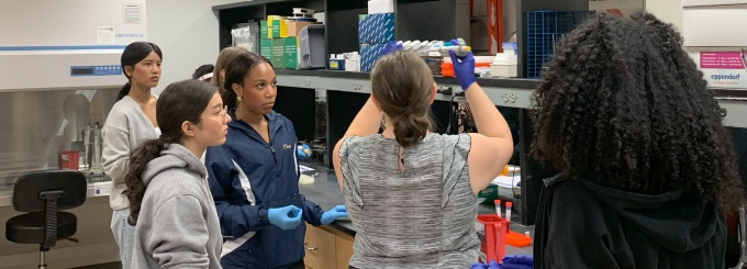Group in a lab observing a demonstration at a workbench with shelves of supplies and equipment. 