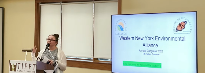Speaker stands at a podium labeled “Tifft Nature Preserve,” addressing an audience with a microphone and notes in hand. A large screen beside the speaker displays “Western New York Environmental Alliance Annual Congress 2026” with logos and a highlighted introduction line. 