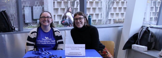 An indoor informational booth for the University at Buffalo School of Public Health and Health Professions. The table is covered with a blue tablecloth featuring the school’s logo and displays brochures, pens, blue lanyards, and a beige tote bag with the same logo. A sign on the table reads “Center for Climate Change and Health Equity” with additional text mentioning “Environmental Health.” Behind the table are two seated individuals, and the background shows large windows, a person sitting outside, and bags placed on the floor. 