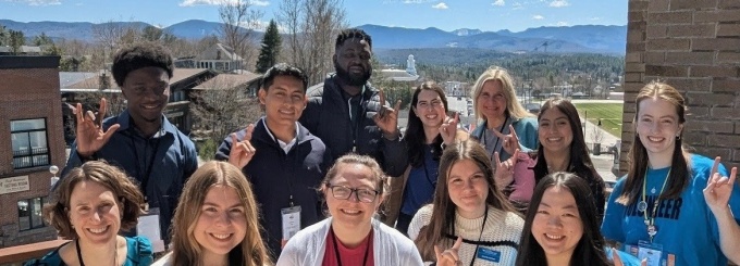Group of people standing close together on an outdoor deck, posing for a photo with mountains and a bright blue sky in the background. Several individuals wear name badges and hold up hand signs, while buildings and trees are visible below the overlook. 