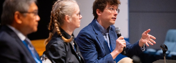 Three panelists sit at a conference table on a stage. One speaker in a blue blazer holds a handheld microphone and gestures while speaking, while two others sit beside them slightly out of focus. A screen and chairs are visible in the background. 