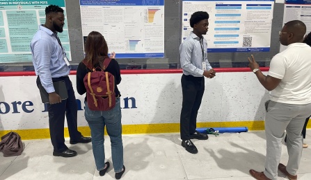 People stand in front of a row of scientific poster presentations displayed along a wall, discussing and examining the materials. One person with a red backpack faces a poster in the center, while another person to the left holds a tablet and looks toward the same display. On the right, two people face each other in conversation, with one gesturing toward a blue poster. The posters contain charts, graphs, and text about public health topics, and are mounted behind clear panels on a rink-style barrier. The setting appears to be an indoor event space with a worn white floor and a red-and-blue backdrop above the posters. 