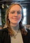 Close-up portrait of a person wearing round, thin-framed glasses and a gray turtleneck with a sun-shaped pendant necklace. The person has shoulder-length light brown hair and is indoors, with soft lighting, plants, and industrial-style ceiling fixtures blurred in the background. 