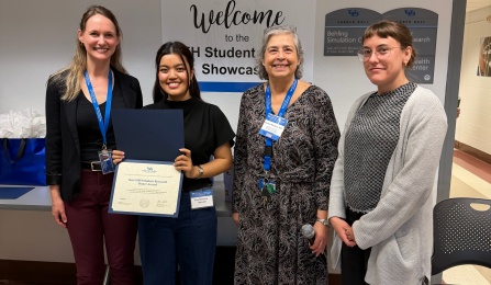 Four people stand in a hallway at a University at Buffalo event, with a sign for the EEH Student Showcase behind them. One person holds an open folder displaying an award certificate while others wear lanyards and stand beside them. 