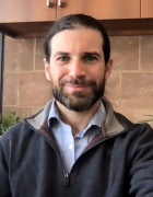 Photograph of the upper torso and head of a white man with dark hair wearing a blue button up and dark fleece jacket. 