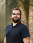 Person with short dark hair, glasses, and a beard, wearing a black polo shirt, standing outdoors in front of a softly blurred background with greenery and a fence; a tattoo is visible on one forearm.
