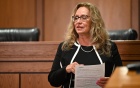 A person standing in a courtroom setting, holding a printed document with highlighted text. The individual is wearing a black outfit with white trim and a pearl necklace. Behind them are wooden panels and two black leather chairs, suggesting a judicial or formal environment. 