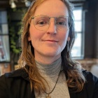 Close-up portrait of a person wearing round, thin-framed glasses and a gray turtleneck with a sun-shaped pendant necklace. The person has shoulder-length light brown hair and is indoors, with soft lighting, plants, and industrial-style ceiling fixtures blurred in the background.