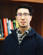Photograph of an Asian man with glasses from the shoulders up in a dark zippered sweater and a white and black patterned shirt. He stands in front of a bookshelf. 