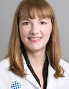 Image of the face and upper torso of a woman with bangs and shoulder length red hair in a white lab coat with a UBMD logo. 