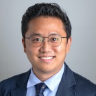 A person wearing a dark suit jacket, light blue collared shirt, and a navy blue tie with small gold patterns, posed against a plain light gray background. 
