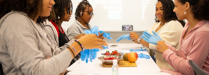 Students seated at a table put on blue food prep gloves. 