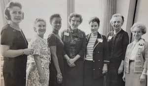 seven women pose for a photo in front of a window. 