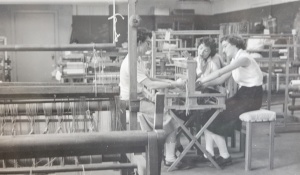 in a large room full of wooden looms, three women sit at a loom. 